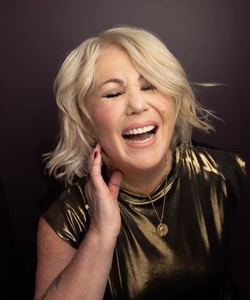 Jann Arden smiling with her eyes shut. She has blonde hair and wears a gold top. 