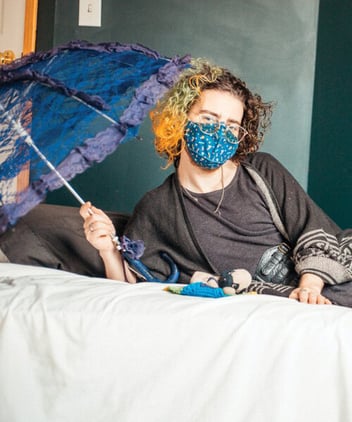 A landscape picture of Oliver, a white-appearing non-binary artist. They are casually leaning onto a mattress on their right elbow with their legs on the ground. A tiny 22 year-old stuffed dog, named Beethoven, lays in front of them on the mattress with a small rainbow blanket. Oliver has a purple lace umbrella in their right hand. They are wearing glasses, a blue bus-print mask, dark green shawl, a grey dress with a brown paisley velvet skirt underneath it, green leg warmers, and heels with cat faces on them. Their curly shoulder length hair is orange and green on one side and brown on the other. There is a softness in this image as Oliver and Beethoven bask in the light from a window together.
