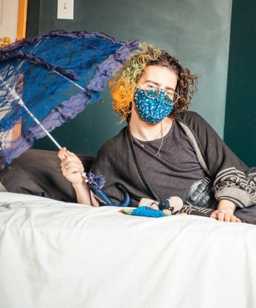 A landscape picture of Oliver, a white-appearing non-binary artist. They are casually leaning onto a mattress on their right elbow with their legs on the ground. A tiny 22 year-old stuffed dog, named Beethoven, lays in front of them on the mattress with a small rainbow blanket. Oliver has a purple lace umbrella in their right hand. They are wearing glasses, a blue bus-print mask, dark green shawl, a grey dress with a brown paisley velvet skirt underneath it, green leg warmers, and heels with cat faces on them. Their curly shoulder length hair is orange and green on one side and brown on the other. There is a softness in this image as Oliver and Beethoven bask in the light from a window together.